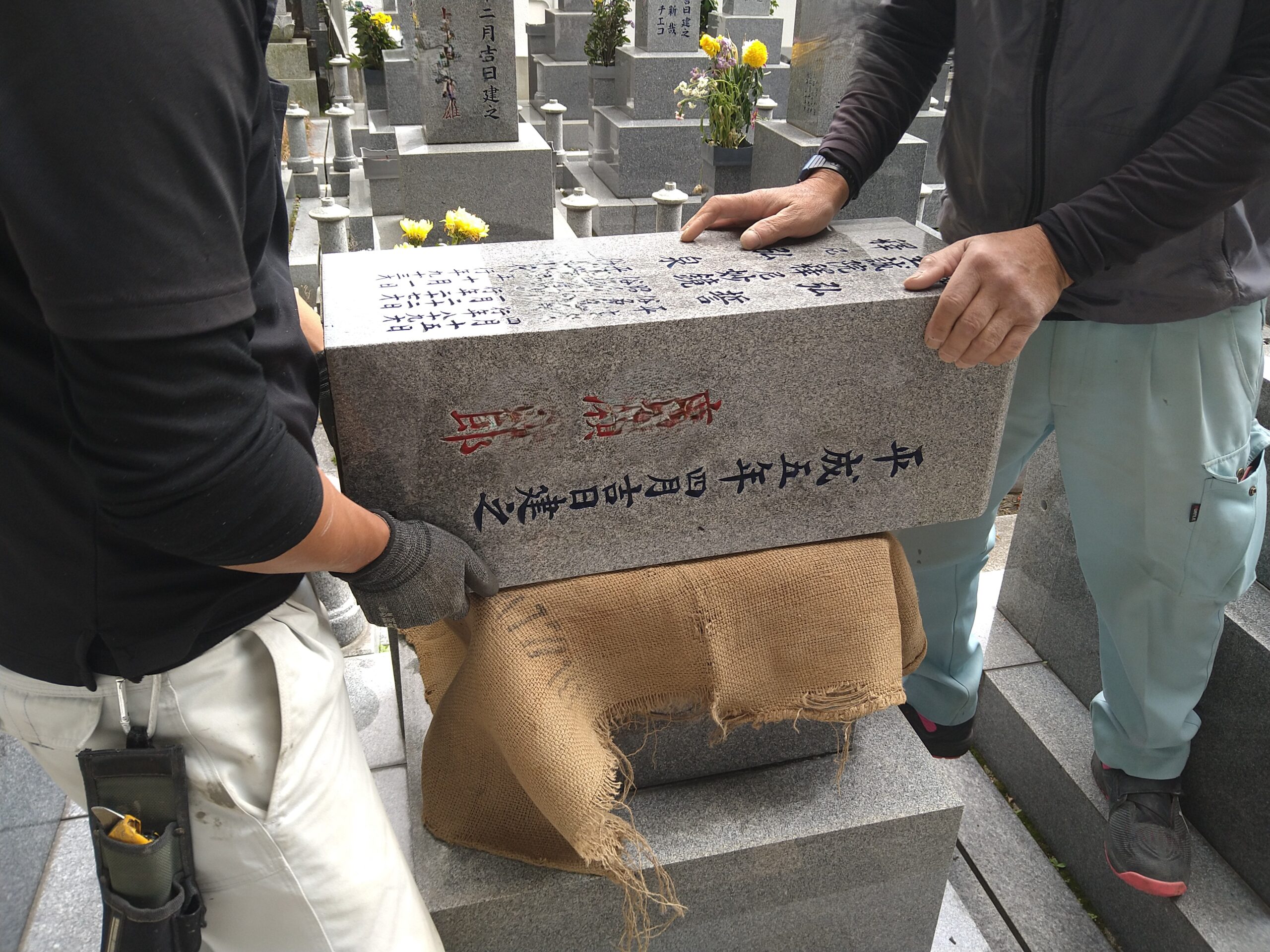 墓所にて二人の職人が重い石材を丁寧に持ち上げ、据え付けている様子